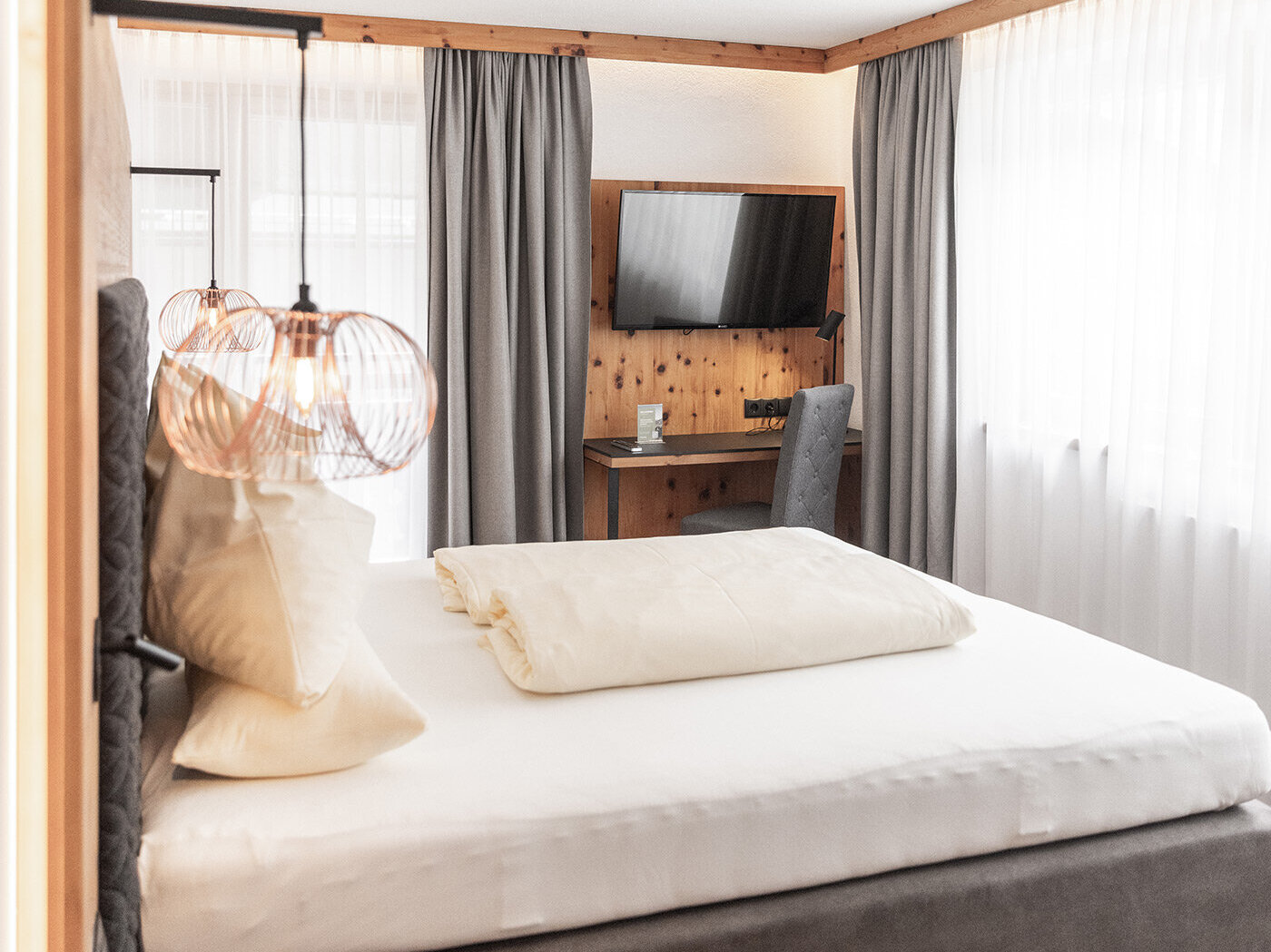 Ein modernes Schlafzimmer mit einem ordentlich gemachten Bett, einem grauen Kopfteil, zwei hängenden Pendelleuchten, einem Schreibtisch mit Stuhl, einem an der Wand montierten Fernseher und großen Fenstern mit grauen Vorhängen, durch die Tageslicht hereinströmt.