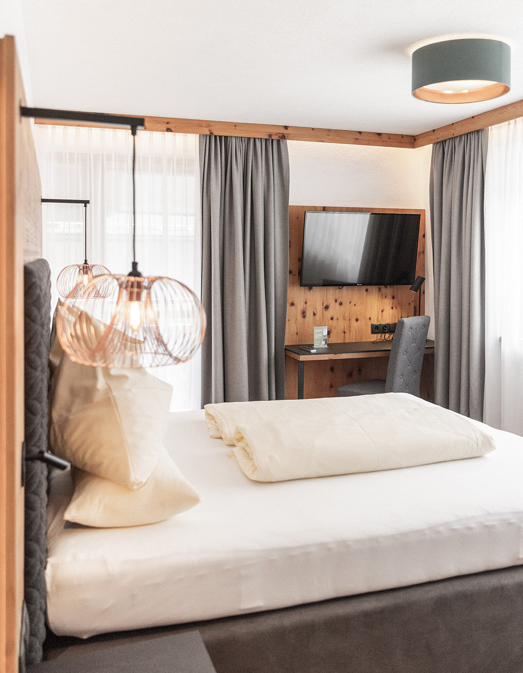 Ein modernes Schlafzimmer mit einem ordentlich gemachten Bett, einem grauen Kopfteil, zwei hängenden Pendelleuchten, einem Schreibtisch mit Stuhl, einem an der Wand montierten Fernseher und großen Fenstern mit grauen Vorhängen, durch die Tageslicht hereinströmt.
