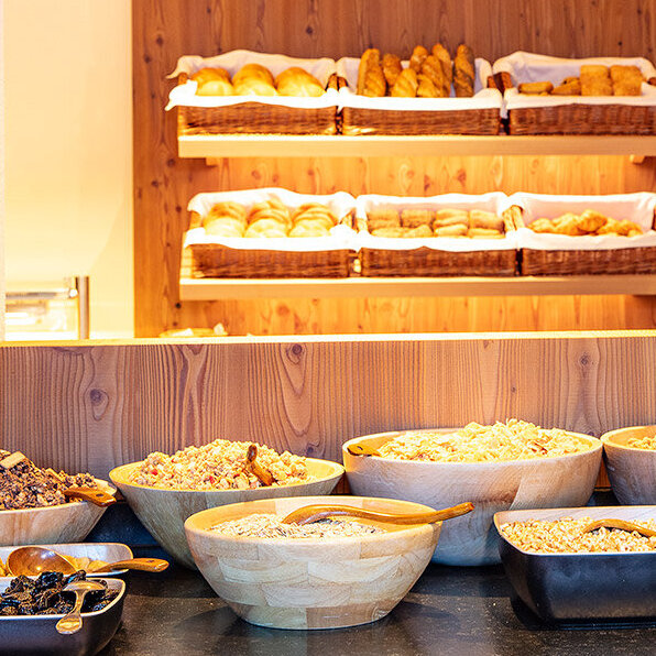 Im Vordergrund ein Frühstücksbuffet mit Holzschalen voller Müsli, Trockenfrüchten und Nüssen, im Hintergrund Regale mit verschiedenen Brotsorten und Gebäck in Körben.