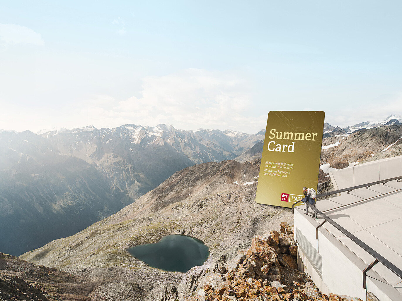 Auf einem Bergplateau sieht man eine große Grafik der goldenen Summer Card mit Blick auf eine malerische Alpenlandschaft mit fernen Gipfeln und einem See; in der Nähe ist eine Person und genießt die Aussicht.