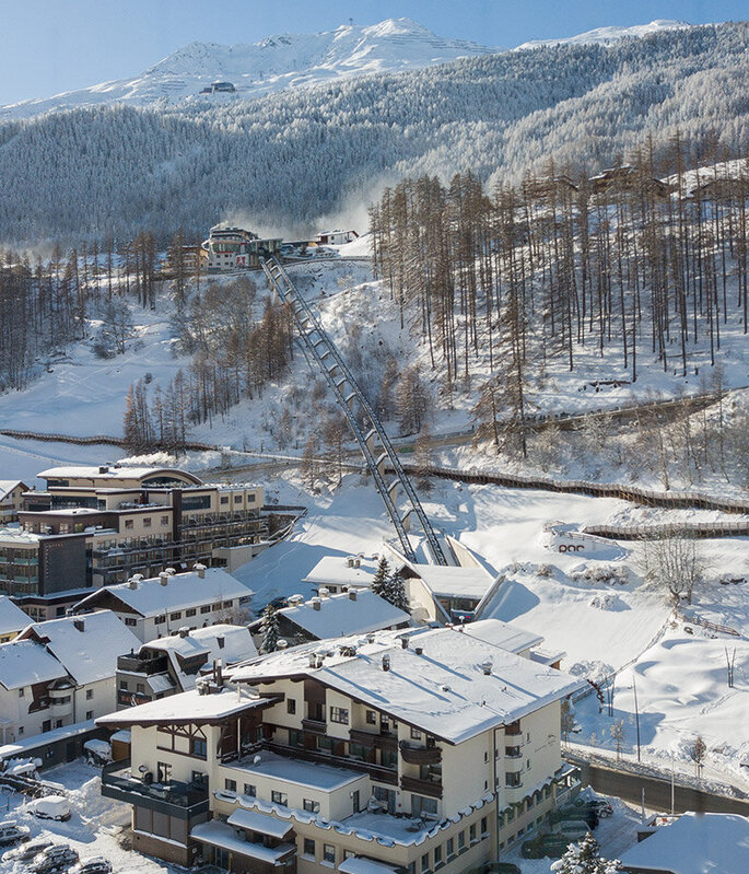 Ein verschneites Bergdorf mit Häusern, geparkten Autos und schneebedeckten Bäumen. Der Zentrum Shuttle führt hinter dem Hotel Hubertus den Hang hinauf, im Hintergrund sind Berge und ein strahlend blauer Himmel zu sehen.