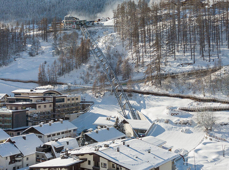 Ein verschneites Bergdorf mit Häusern, geparkten Autos und schneebedeckten Bäumen. Der Zentrum Shuttle führt hinter dem Hotel Hubertus den Hang hinauf, im Hintergrund sind Berge und ein strahlend blauer Himmel zu sehen.
