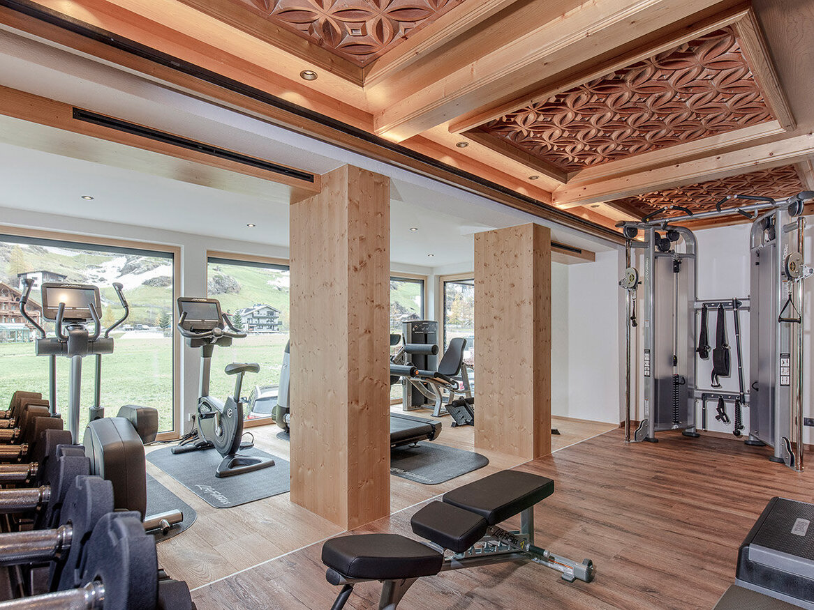 Ein modernes Fitnessstudio mit Hanteln, Trainingsbänken, Cardiogeräten und Krafttrainingsgeräten. Große Fenster sorgen für Tageslicht und bieten einen malerischen Blick ins Freie. Die kunstvoll gestaltete Holzdecke ist gut zu sehen.