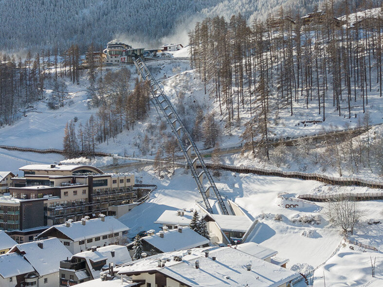Ein verschneites Bergdorf mit Häusern, geparkten Autos und schneebedeckten Bäumen. Der Zentrum Shuttle führt hinter dem Hotel Hubertus den Hang hinauf, im Hintergrund sind Berge und ein strahlend blauer Himmel zu sehen.