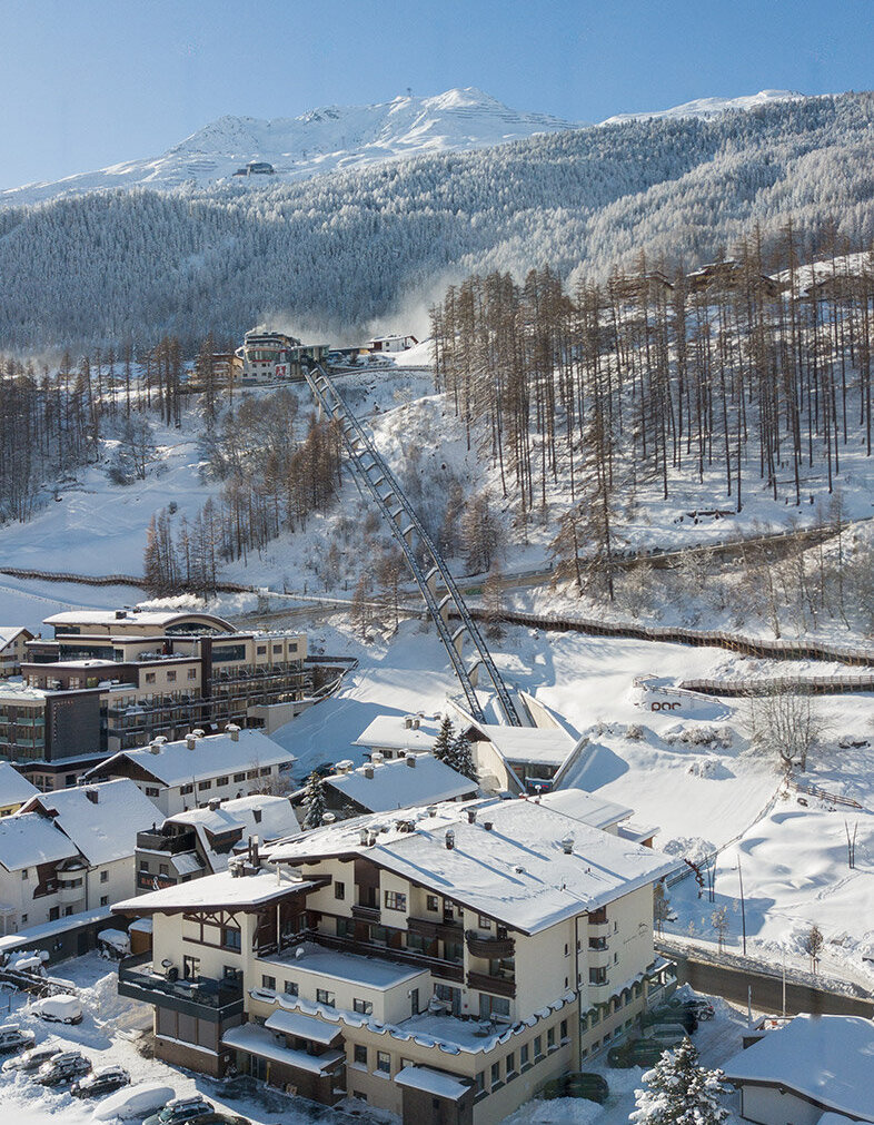 Ein verschneites Bergdorf mit Häusern, geparkten Autos und schneebedeckten Bäumen. Der Zentrum Shuttle führt hinter dem Hotel Hubertus den Hang hinauf, im Hintergrund sind Berge und ein strahlend blauer Himmel zu sehen.