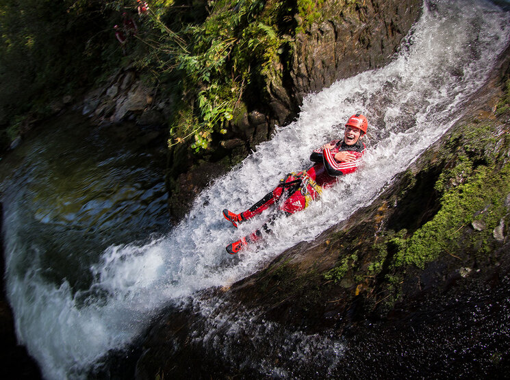 Eine Person mit Helm und Outdoor-Ausrüstung rutscht an einem felsigen Hang einen kleinen Wasserfall hinunter, lächelt und verschränkt die Arme, umgeben von Grün und fließendem Wasser.
