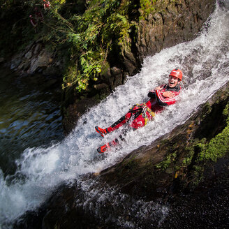 Eine Person mit Helm und Outdoor-Ausrüstung rutscht an einem felsigen Hang einen kleinen Wasserfall hinunter, lächelt und verschränkt die Arme, umgeben von Grün und fließendem Wasser.