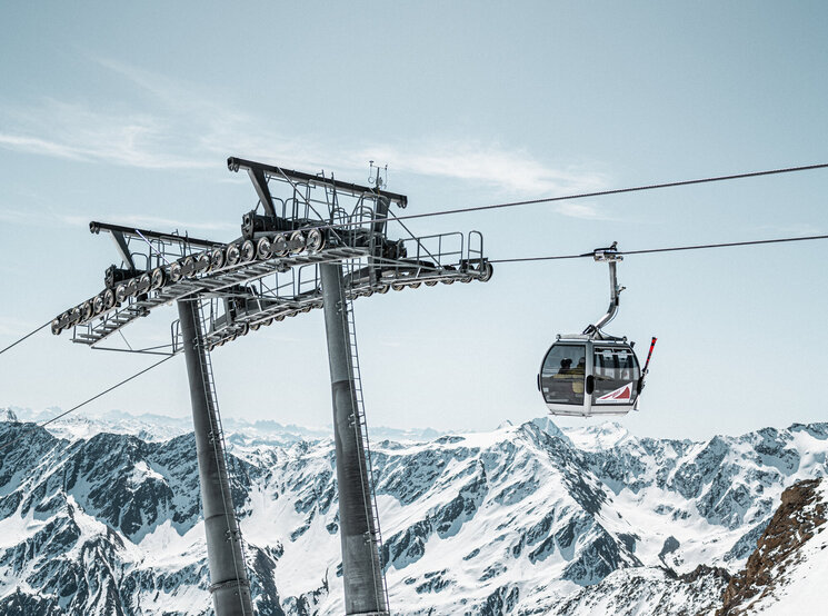 Eine Seilbahnkabine fährt unter klarem Himmel über verschneite Berge, gestützt von hohen Metallmasten. Die Berglandschaft erstreckt sich bis in die Ferne.