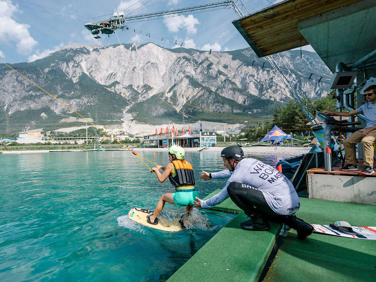 Eine Person mit Helm und Schwimmweste beginnt an einem Steg mit dem Wakeboarden an einem Seil, im Hintergrund sind Berge und Gebäude zu sehen. Mehrere Menschen schauen in der Nähe zu.