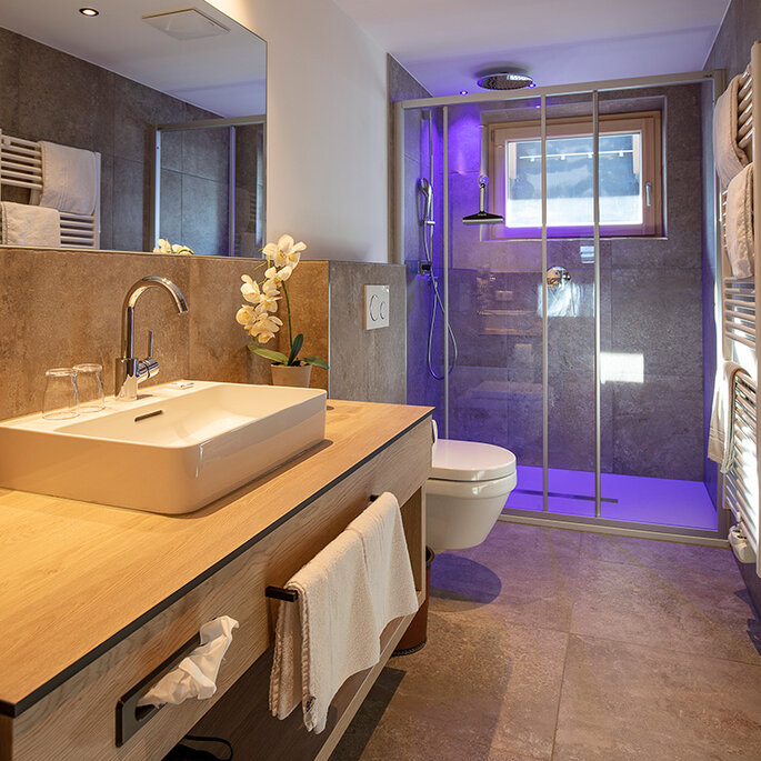 Modernes Badezimmer mit einem großen Spiegel, einem Aufsatzwaschbecken auf einem Waschtisch aus Holz, einer begehbaren Dusche mit Glastüren und violetter Beleuchtung im Duschbereich. Neutrale Fliesen an Boden und Wänden.