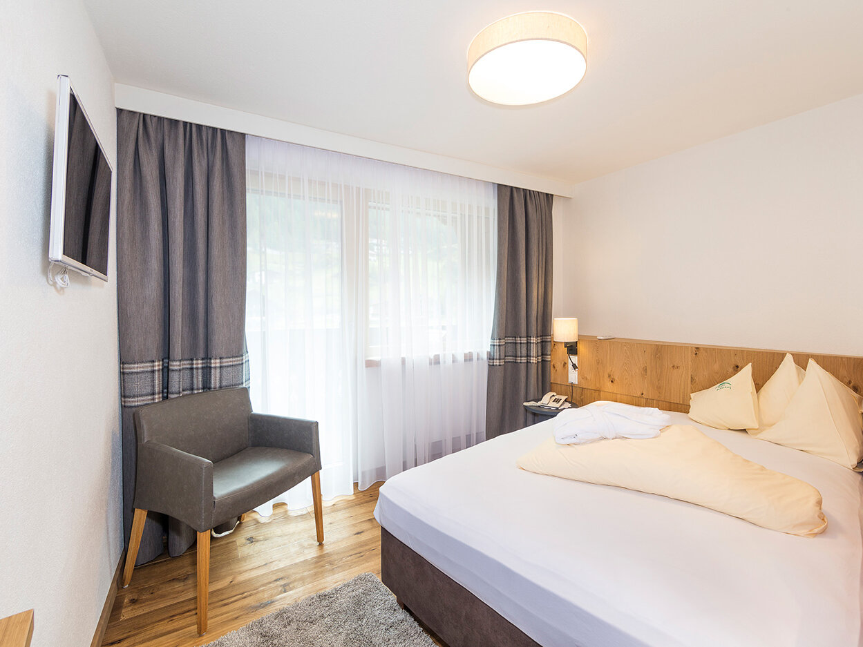 Modernes Hotelzimmer mit einem Doppelbett, einem grauen Sessel, einem an der Wand montierten Fernseher, einem Holzboden und einem großen Fenster, das mit transparenten Vorhängen und dunklen Gardinen verdeckt ist. Auf dem Bett liegt ein gefaltetes Handtuch.