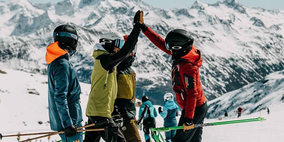 Drei Skifahrer in Winterkleidung geben sich auf einem verschneiten Berghang ein High-Five, im Hintergrund sind schneebedeckte Gipfel und weitere Skifahrer zu sehen, über ihnen ein klarer Himmel.
