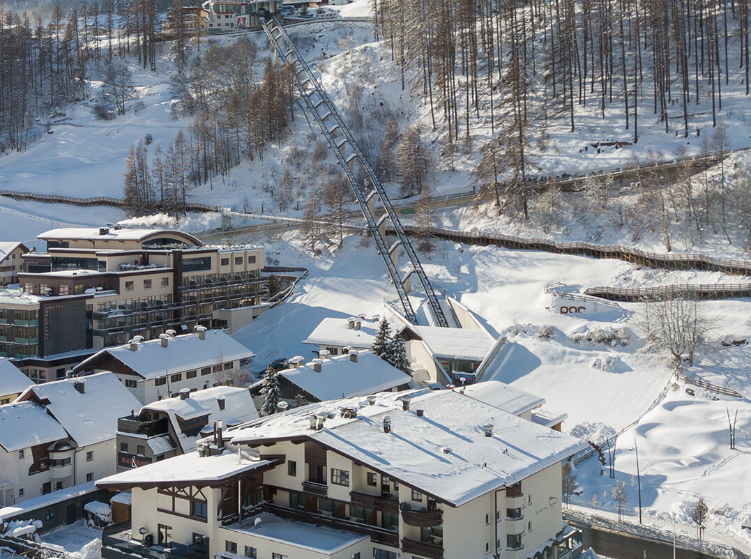 Ein verschneites Bergdorf mit Häusern, geparkten Autos und schneebedeckten Bäumen. Der Zentrum Shuttle führt hinter dem Hotel Hubertus den Hang hinauf, im Hintergrund sind Berge und ein strahlend blauer Himmel zu sehen.