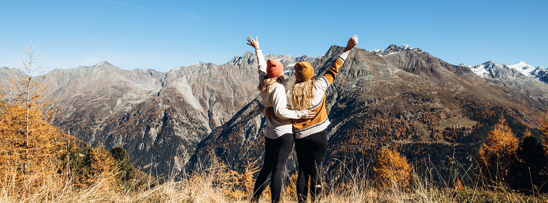Zwei Menschen stehen Seite an Seite mit erhobenen Armen und blicken unter einem strahlend blauen Himmel auf eine malerische Berglandschaft. Im Vordergrund sind herbstlich gefärbte Bäume zu sehen.