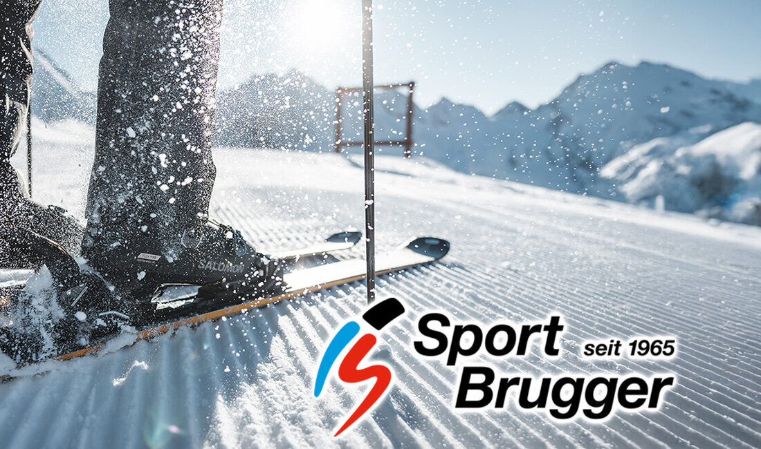 Nahaufnahme der Skischuhe eines Skifahrers auf Skiern im Neuschnee, wobei Schnee hochspritzt. Im Hintergrund sind Berge und Sonnenlicht zu sehen. Unten rechts ist das Logo „Sport Brugger seit 1965“ zu erkennen.