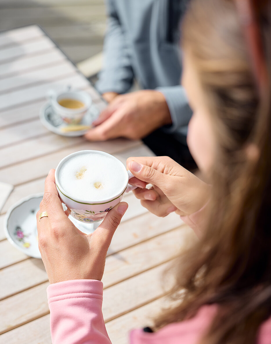 Eine Frau in einem rosa Oberteil sitzt mit einer Tasse Cappuccino an einem Tisch im Freien zusammen mit einer anderen Person, die ebenfalls eine Tasse Kaffee vor sich hat.