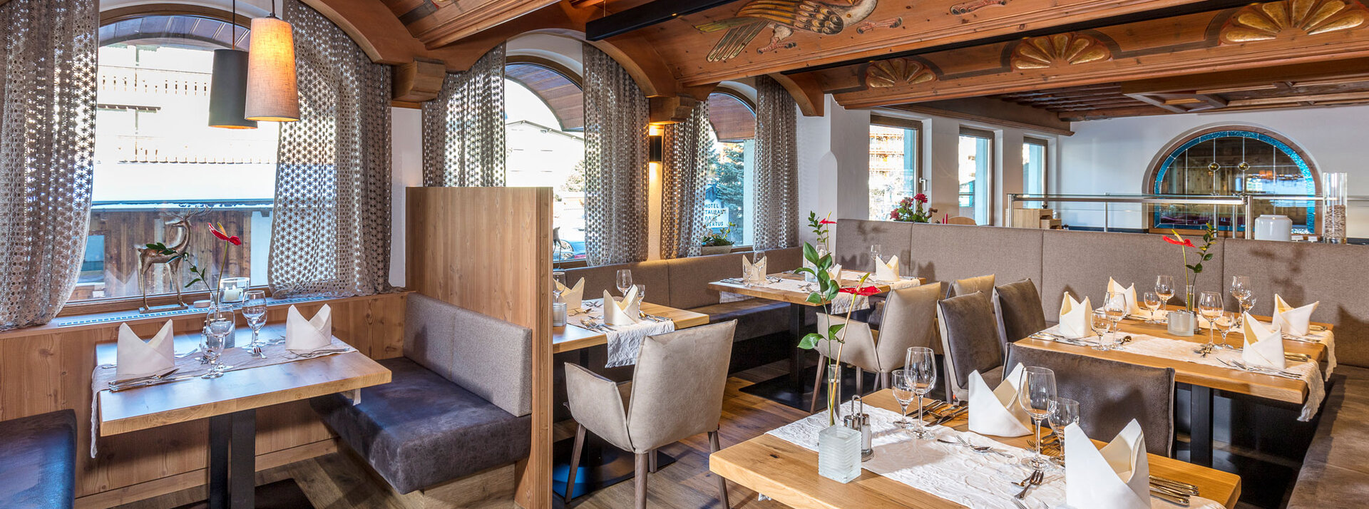 Ein gemütliches Restaurantinterieur mit gedeckten Holztischen, gepolsterten Stühlen, Bogenfenstern und dekorativen Holzverzierungen an der Decke. Natürliches Licht erhellt den eleganten und einladenden Raum.