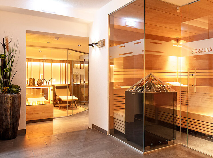 Ein modernes Spa-Interieur mit einer Sauna mit Glaswänden, die mit Holzbänken, einer zentralen Heizung und warmer Beleuchtung ausgestattet ist. Im angrenzenden Raum befinden sich Topfpflanzen und Sitzecken.