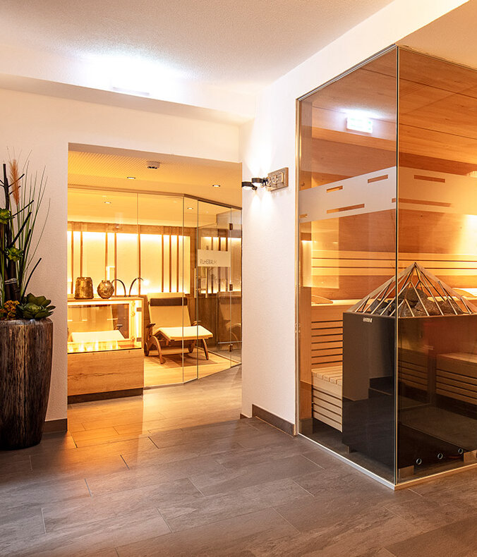 Ein modernes Spa-Interieur mit einer Sauna mit Glaswänden, die mit Holzbänken, einer zentralen Heizung und warmer Beleuchtung ausgestattet ist. Im angrenzenden Raum befinden sich Topfpflanzen und Sitzecken.