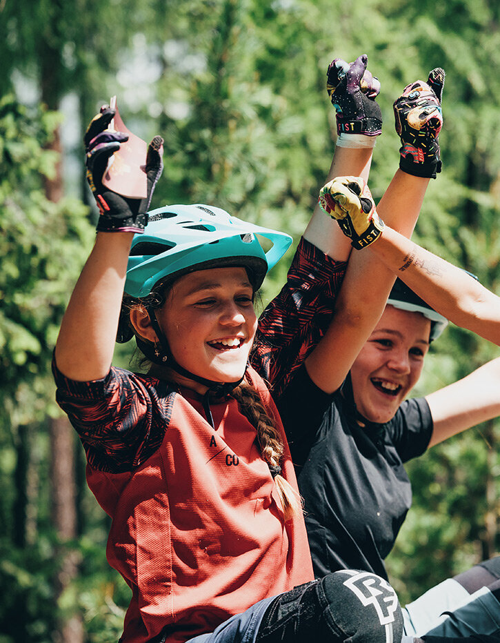 Drei Kinder mit Fahrradhelmen und Fahrradbekleidung sitzen im Freien, lächeln und strecken vor Begeisterung die Arme in die Höhe, im Hintergrund ist ein Wald mit grünen Bäumen zu sehen.