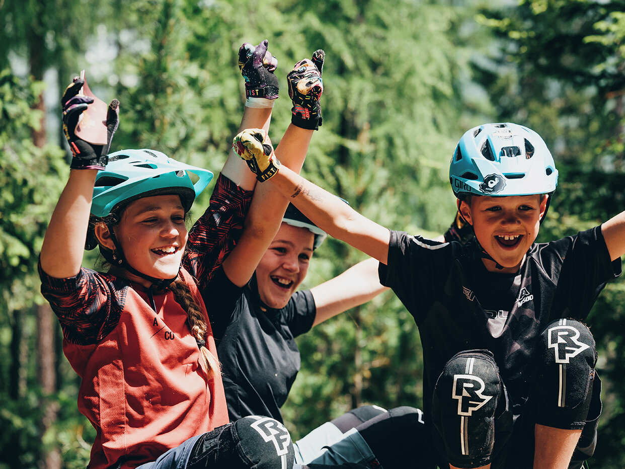 Drei Kinder mit Fahrradhelmen und Fahrradbekleidung sitzen im Freien, lächeln und strecken vor Begeisterung die Arme in die Höhe, im Hintergrund ist ein Wald mit grünen Bäumen zu sehen.