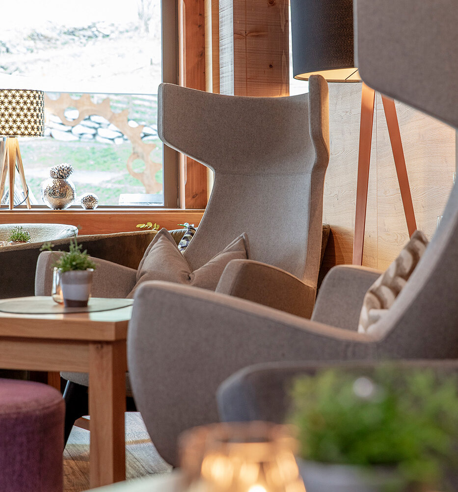 Gemütliche Lounge mit modernen Stühlen mit hoher Rückenlehne, kleinen Topfpflanzen auf Holztischen, weichen Kissen, einer Stehlampe und einer Kerze im Vordergrund. Große Fenster lassen Tageslicht herein.