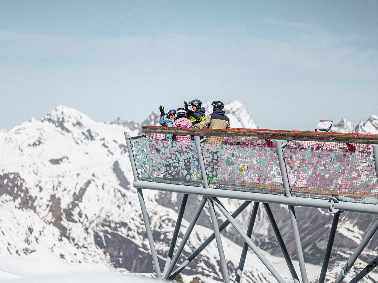 Eine Gruppe von Menschen in Winterkleidung steht auf einer Aussichtsplattform mit Blick auf schneebedeckte Berge unter einem klaren Himmel.