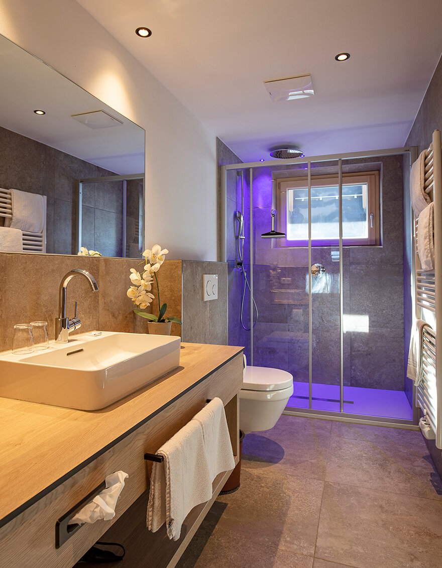 Modernes Badezimmer mit einem Waschtisch aus Holz, einem weißen Waschbecken, einem Spiegel, einem Handtuch und einer Glasduschkabine mit violetter Beleuchtung. Neutrale Farbtöne, geflieste Wände und Böden sowie ein Fenster über der Dusche sorgen für natürliches Licht.