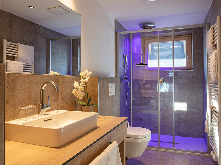 Modernes Badezimmer mit einem Waschtisch aus Holz, einem weißen Waschbecken, einem Spiegel, einem Handtuch und einer Glasduschkabine mit violetter Beleuchtung. Neutrale Farbtöne, geflieste Wände und Böden sowie ein Fenster über der Dusche sorgen für natürliches Licht.