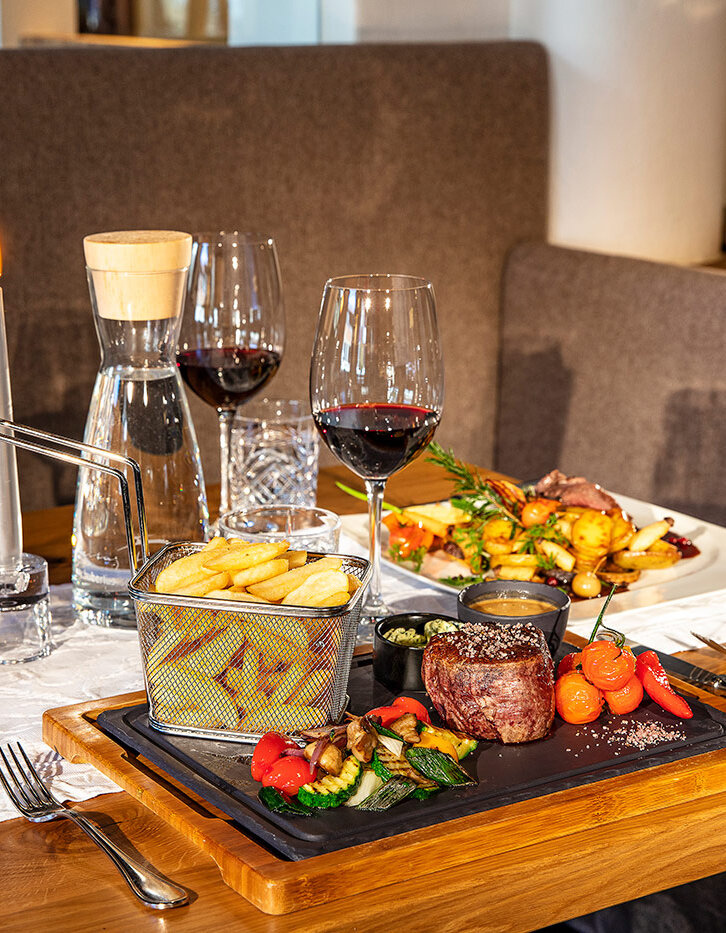 Ein für zwei Personen gedeckter Restauranttisch mit Rotwein, Steak, Pommes frites, Gemüse und einer Blumenvase. Der Tisch ist mit einer brennenden Kerze und einer Wasserkaraffe geschmückt, was eine gemütliche Atmosphäre beim Essen schafft.
