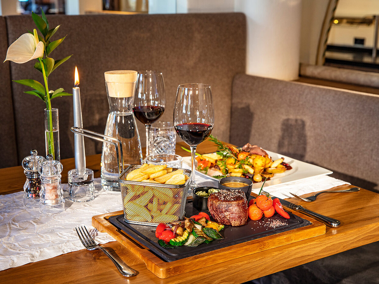 Ein für zwei Personen gedeckter Restauranttisch mit Rotwein, Steak, Pommes frites, Gemüse und einer Blumenvase. Der Tisch ist mit einer brennenden Kerze und einer Wasserkaraffe geschmückt, was eine gemütliche Atmosphäre beim Essen schafft.