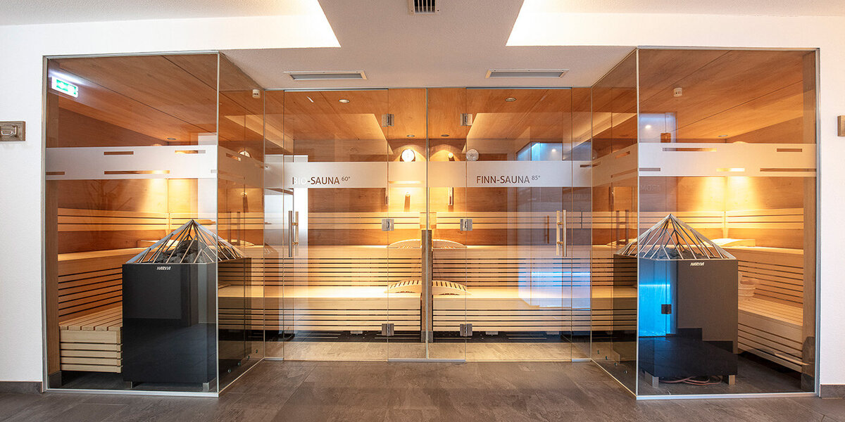 Eine moderne finnische Sauna mit Holzbänken und Glaswänden, die über zwei große Öfen in der Mitte verfügt und durch eine warme Beleuchtung eine entspannende Atmosphäre schafft.