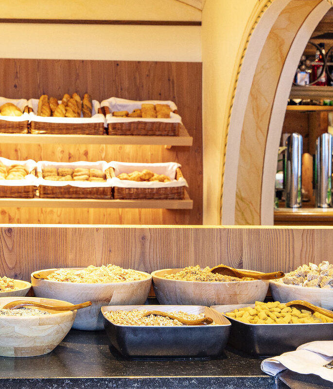 Im Vordergrund ein Frühstücksbuffet mit Holzschalen voller Müsli, Trockenfrüchten und Nüssen, im Hintergrund Regale mit verschiedenen Brotsorten und Gebäck in Körben.