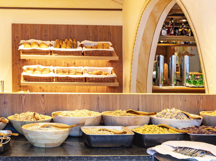 Im Vordergrund ein Frühstücksbuffet mit Holzschalen voller Müsli, Trockenfrüchten und Nüssen, im Hintergrund Regale mit verschiedenen Brotsorten und Gebäck in Körben.