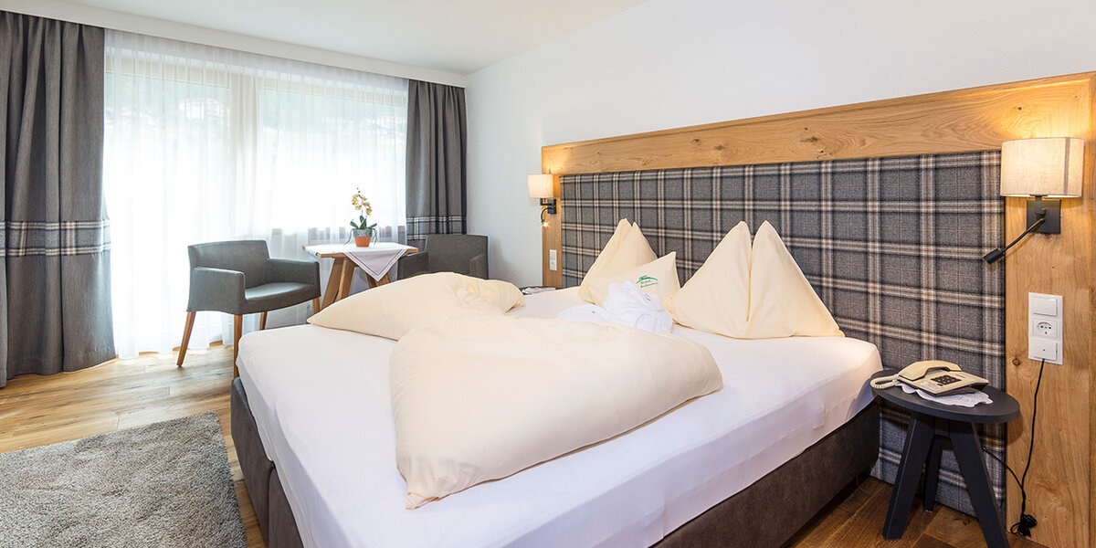 Ein gemütliches Hotelzimmer mit einem großen Bett, einem karierten Kopfteil, zwei Sesseln am Fenster, einem kleinen runden Tisch mit Blumen, einem Holzboden und moderner Beleuchtung.