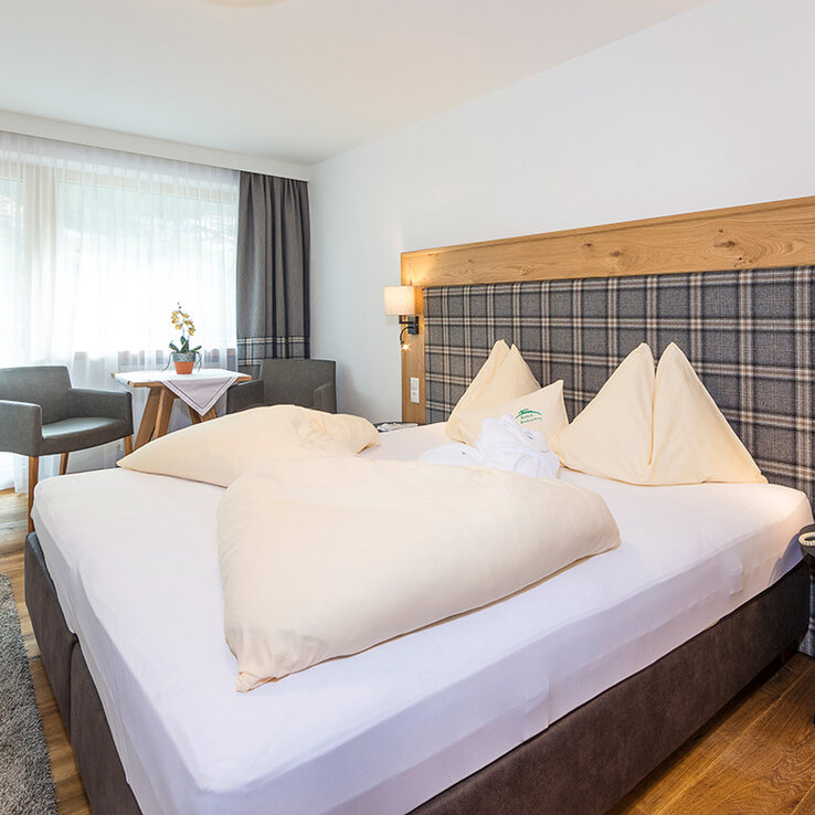 Ein gemütliches Hotelzimmer mit einem großen Bett, einem karierten Kopfteil, zwei Sesseln am Fenster, einem kleinen runden Tisch mit Blumen, einem Holzboden und moderner Beleuchtung.