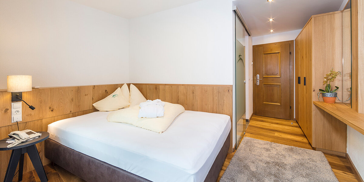 Ein modernes Hotelzimmer mit einem ordentlich gemachten Bett, Holzvertäfelung, einem Nachttisch, einem Einbauschrank, einer Tür und einer kleinen Pflanze auf einem Regal. Das Zimmer besticht durch sanfte Beleuchtung und ein gemütliches, minimalistisches Design.