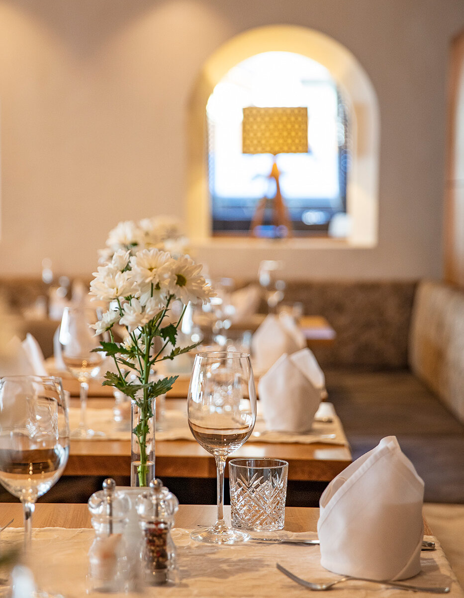 Ein elegant gedeckter Restauranttisch mit weißen Servietten, Weingläsern, einem Kristallglas und einer Vase mit weißen Blumen. Sanfte Beleuchtung und eine Lampe im Hintergrund sorgen für eine warme, einladende Atmosphäre.