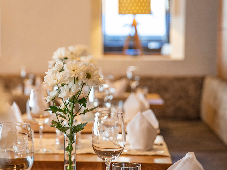 Ein elegant gedeckter Restauranttisch mit weißen Servietten, Weingläsern, einem Kristallglas und einer Vase mit weißen Blumen. Sanfte Beleuchtung und eine Lampe im Hintergrund sorgen für eine warme, einladende Atmosphäre.