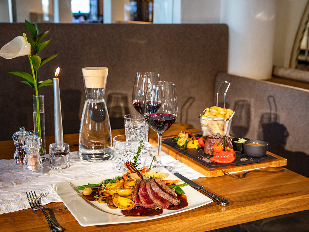 Ein Restauranttisch, gedeckt mit Wein, Wasser, einer Kerze und zwei Tellern mit Gourmetgerichten, darunter Aufschnitt, Kartoffeln, Gemüse, Pommes frites und Soßen, dazu elegantes Geschirr und eine weiße Serviette.