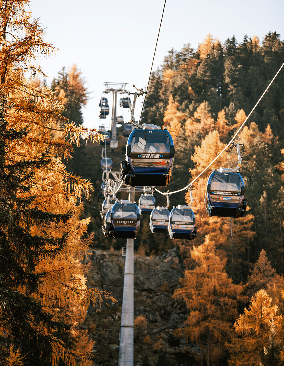 Mehrere blaue Gondelbahnen fahren an einem Seil entlang auf und ab, umgeben von herbstlichen Bäumen mit goldenem Laub und vor dem Hintergrund bewaldeter Berge unter einem klaren Himmel.