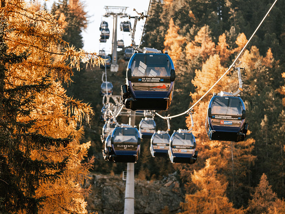 Mehrere blaue Gondelbahnen fahren an einem Seil entlang auf und ab, umgeben von herbstlichen Bäumen mit goldenem Laub und vor dem Hintergrund bewaldeter Berge unter einem klaren Himmel.