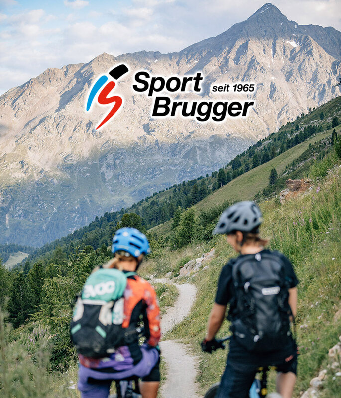 Zwei Mountainbiker fahren auf einem schmalen Pfad durch grüne Hügel, im Hintergrund sind Berge zu sehen. Oben im Bild ist das Logo „Sport Brugger seit 1965“ zu erkennen.