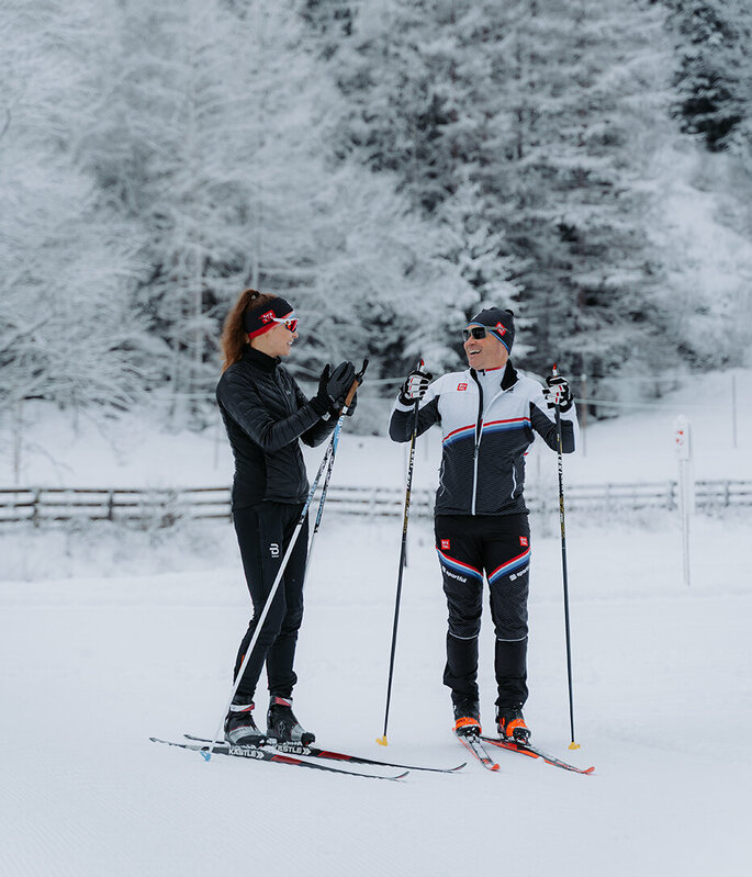 Zwei Personen in Wintersportkleidung sind auf einer verschneiten Loipe beim Langlaufen, umgeben von schneebedeckten Bäumen. Sie stehen da und unterhalten sich, halten ihre Skistöcke in den Händen, im Hintergrund ist ein Holzzaun zu sehen.