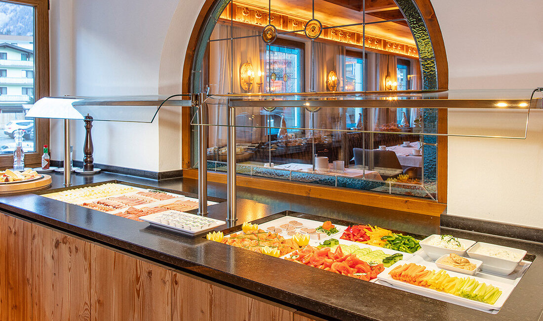 Auf einer Holztheke in einem hellen Raum mit großen Fenstern, der einen gemütlichen Essbereich widerspiegelt, steht ein Buffet mit Platten mit Aufschnitt, Käse, Gemüse und Dips.