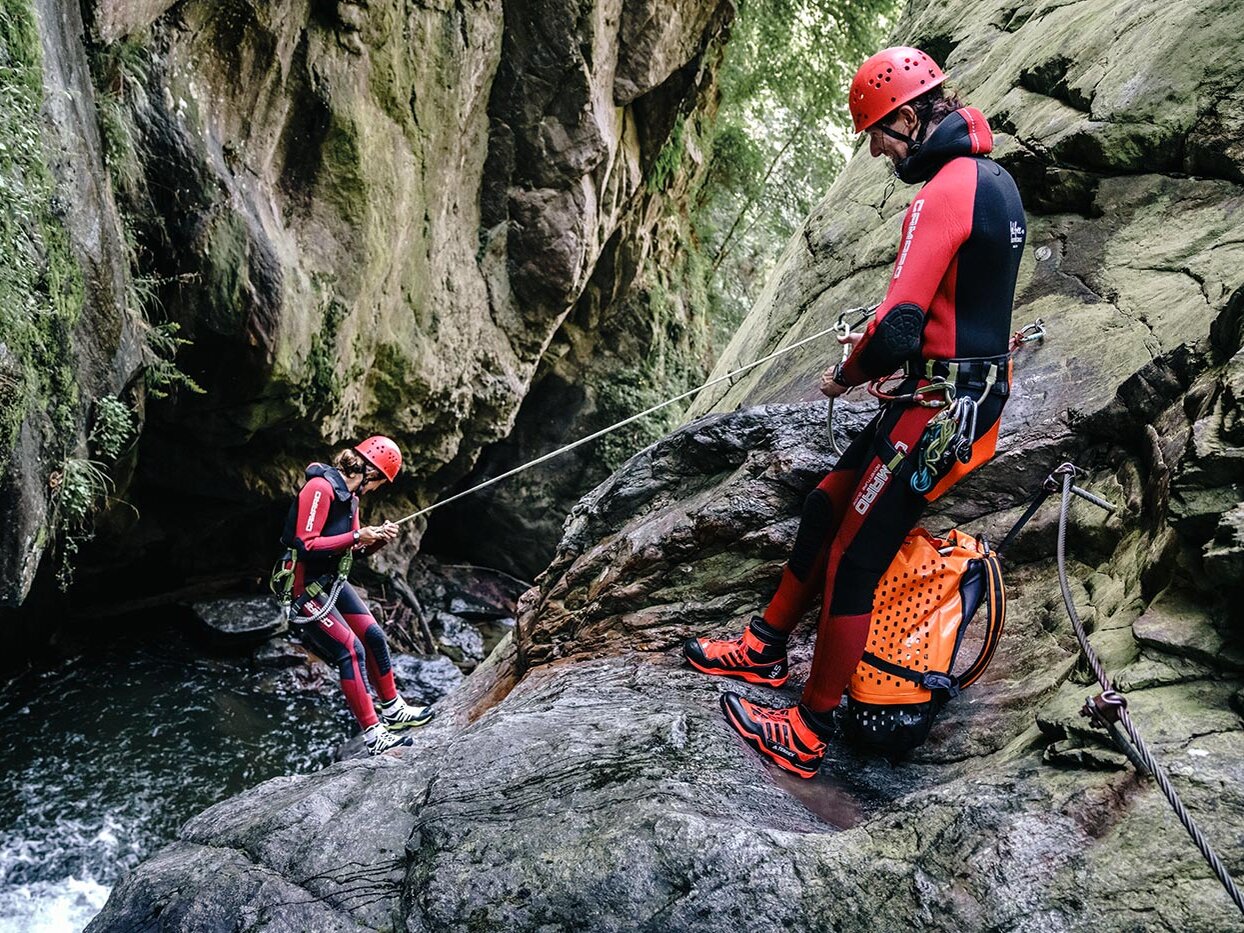 Zwei Personen in roten Neoprenanzügen und Helmen seilen sich an einer felsigen, moosbewachsenen Canyonwand ab; eine von ihnen steigt in Richtung Wasser hinab, während die andere auf einem Felsvorsprung steht und das Seil festhält.