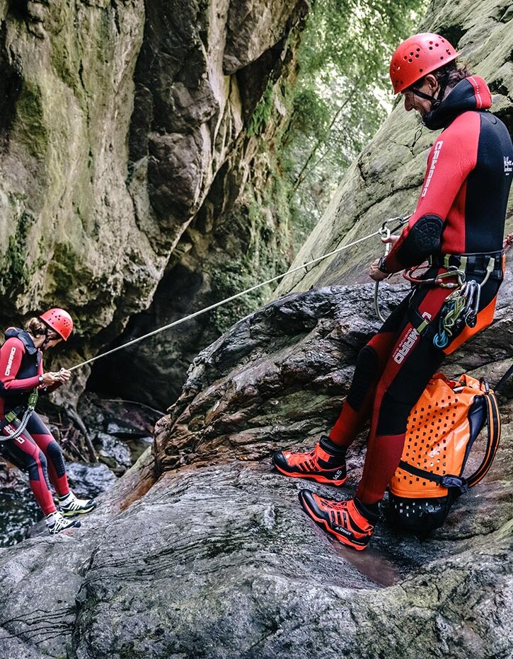 Zwei Personen in roten Neoprenanzügen und Helmen seilen sich an einer felsigen, moosbewachsenen Canyonwand ab; eine von ihnen steigt in Richtung Wasser hinab, während die andere auf einem Felsvorsprung steht und das Seil festhält.