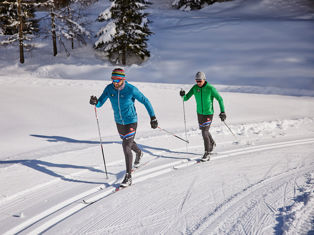 Zwei Menschen fahren auf einer verschneiten Loipe, die von Bäumen gesäumt ist, Langlauf. Beide tragen bunte Jacken und Mützen und gleiten Seite an Seite unter einem strahlenden Winterhimmel dahin.