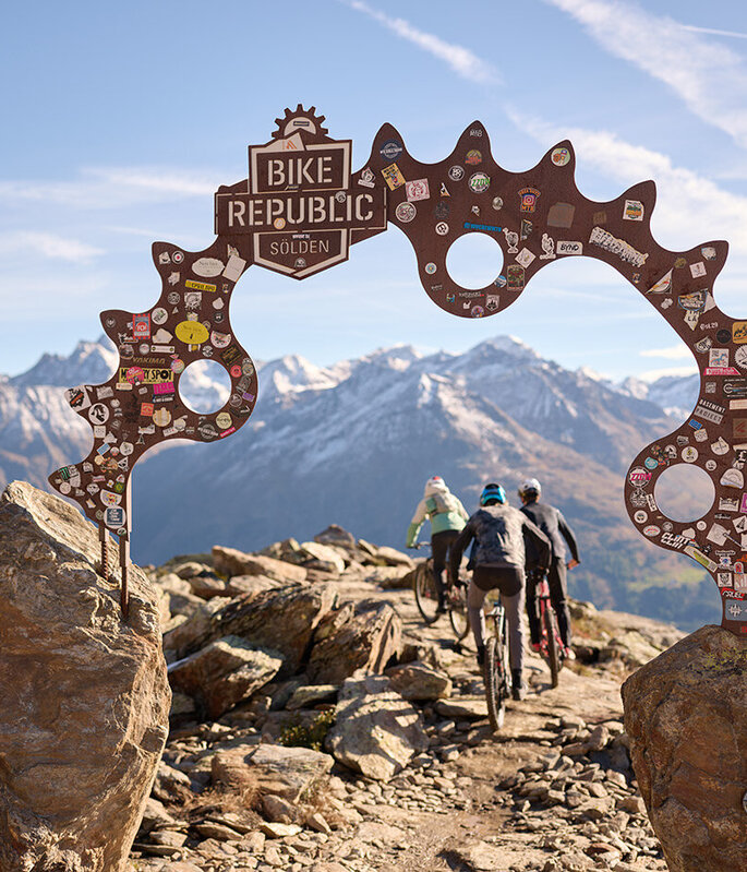 Drei Mountainbiker fahren unter einem Metallbogen mit der Aufschrift „Bike Republic Sölden“ hindurch, umgeben von malerischen schneebedeckten Bergen und einem strahlend blauen Himmel. Der Bogen ist mit Aufklebern verziert.