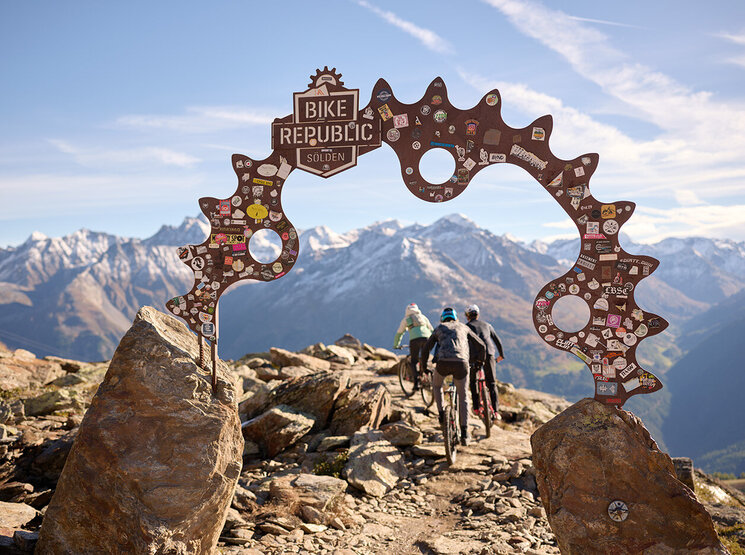 Drei Mountainbiker fahren unter einem Metallbogen mit der Aufschrift „Bike Republic Sölden“ hindurch, umgeben von malerischen schneebedeckten Bergen und einem strahlend blauen Himmel. Der Bogen ist mit Aufklebern verziert.